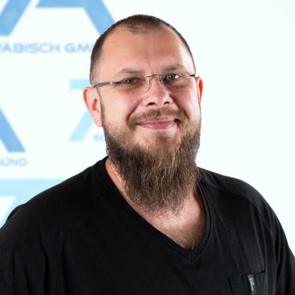 Ein lächelnder Mann mit langem Bart und Brille, der ein schwarzes Hemd trägt, steht vor einem weißen Hintergrund mit blauen geometrischen Formen und teilweise sichtbarem Text.