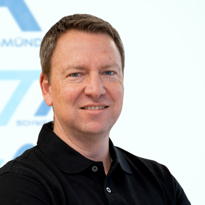 Ein Mann mit kurzen braunen Haaren, der ein schwarzes Polohemd trägt, lächelt in die Kamera. Der Hintergrund ist weiß, darauf sind teilweise blaue Schriftzüge und Formen zu erkennen.
