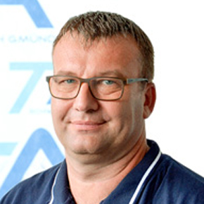 Ein Mann mit kurzen Haaren und Brille, der ein marineblaues Hemd mit Kragen trägt, lächelt in die Kamera. Der Hintergrund ist weiß, darauf sind blauer Text und teilweise erkennbare Formen zu sehen.