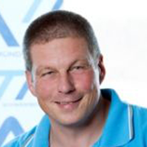 Ein Mann mit kurzen braunen Haaren, der ein hellblaues Hemd mit Kragen trägt, lächelt vor einem unscharfen Hintergrund mit blauen und weißen Mustern in die Kamera.